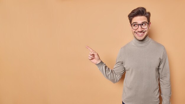 Place your advertisement here Handsome satisfied young European man points at blank space shows place for your promo wears transparent glasses and turtleneck isolated over brown background