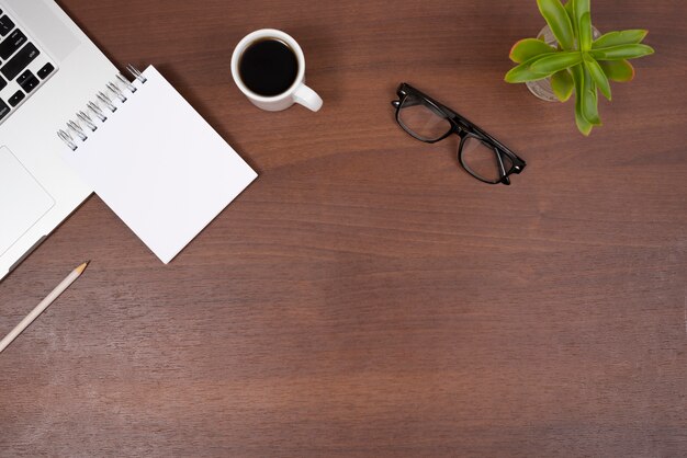 Plant; eye glasses; tea; blank spiral notepad and pencil with laptop on wooden desk