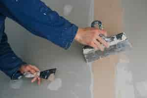 Free photo plasterer man works plastering two trowels on plasterboard in blue uniform