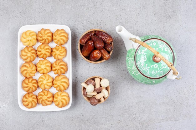 Plate of biscuits next to ornate teapot and bowls of dates and mushroom chocolates on marble background. High quality photo