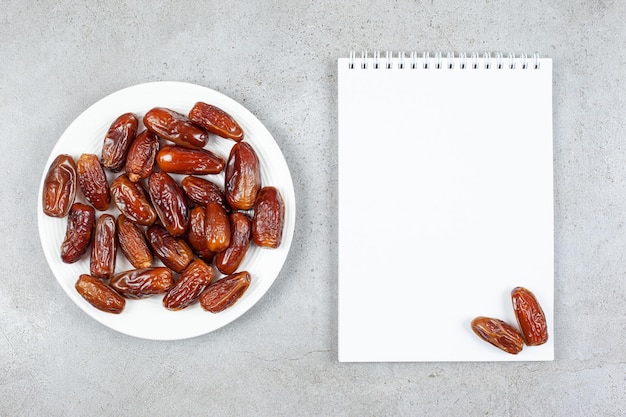 Free photo a plate of fresh dates next to a notebook with two dates on it on marble background.