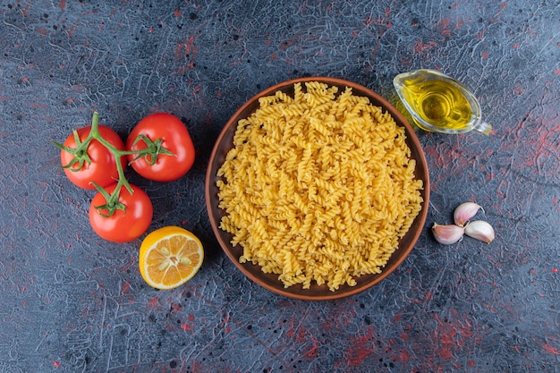 A plate of uncooked spiral pasta with oil and fresh red tomatoes on a dark surface .