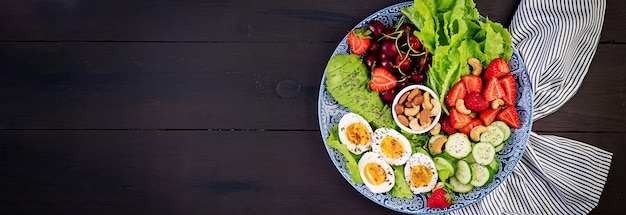 Free Photo plate with a paleo diet food. boiled eggs, avocado, cucumber, nuts, cherry and strawberries. paleo breakfast. top view