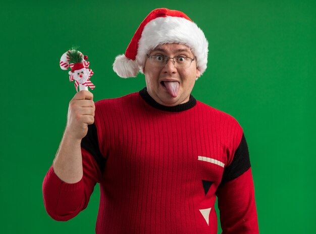 Playful adult man wearing glasses and santa hat holding candy cane ornament showing tongue isolated on green background