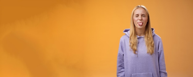 Free photo playful amused charismatic young funny blond girl having fun close eyes showing tongue rebellious fool around unbothered enjoying carefree sunny spring days standing orange background