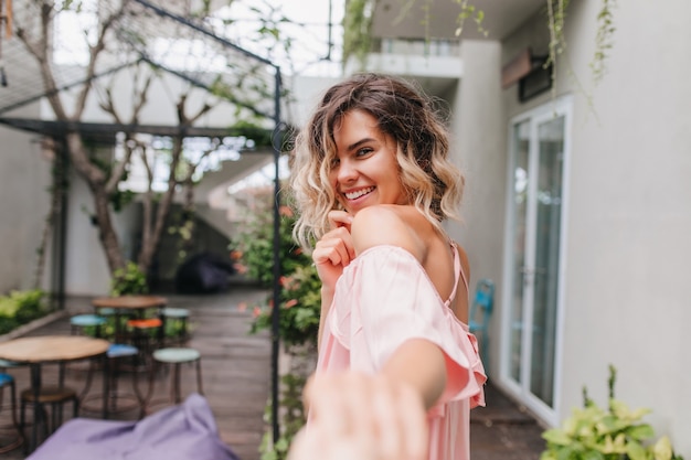 Playful blonde girl looking over shoulder with shy smile. Outdoor photo of chilling caucasian female model having fun in street cafe.