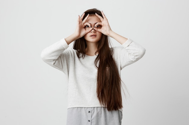 Playful excited teenage female with dark long hair showing Ok gestures with both hands, pretending to wear spectacles, astonished to see something amazing. Emotions, body language and gestures