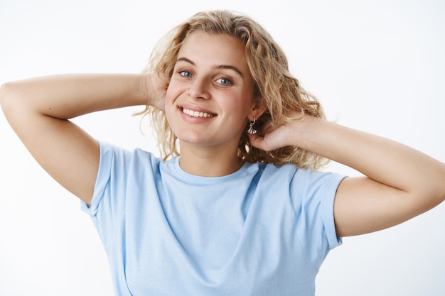 Free Photo playful, feminine attractive european woman with short blond curly hairstyle holding hands behind head as combing hair smiling joyfully and gazing at camera carefree and relaxed over white wall