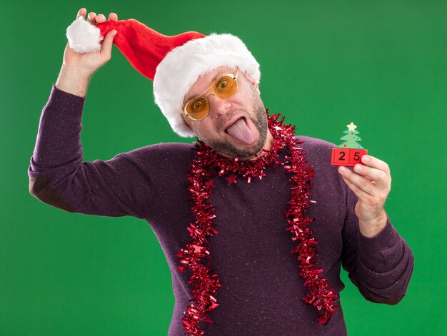 Playful middle-aged man wearing santa hat and tinsel garland around neck with glasses holding christmas tree toy with date