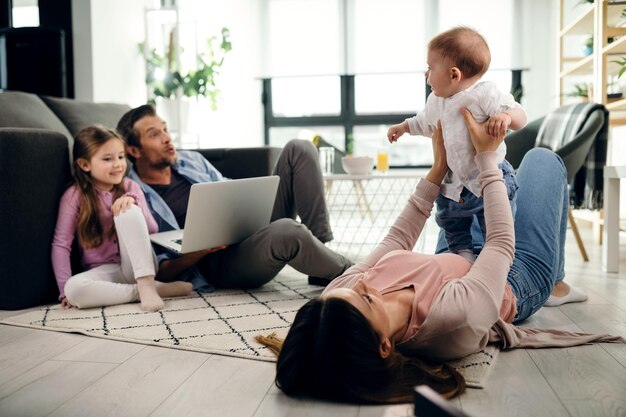 Playful mother holding her baby son while lying on the floor in the living room Father and daughter are using laptop