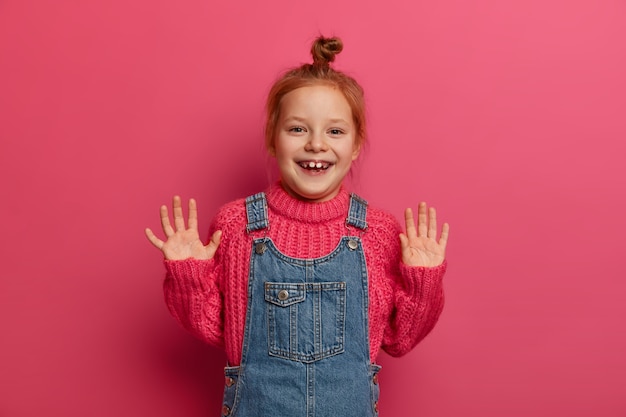 Free photo playful positive girl with ginger hair combed in bun, raises palms and has good mood, poses for family photo in , wears knitted sweater and sarafan, has joyful expression isolated over rosy wall