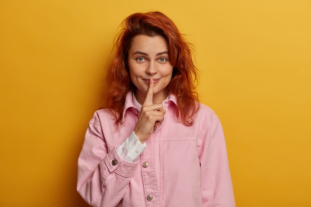 Free Photo playful redhead pretty woman with satisfied secret look, makes hush gesture, gossips with friend, dressed in rosy denim jacket