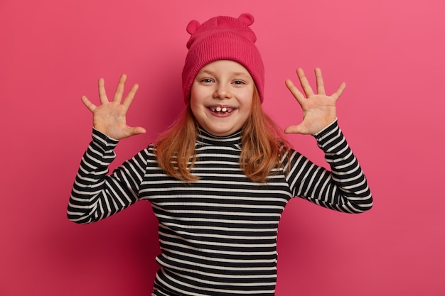 Playful small cute girl raises palms, feels happiness, wears funny hat with ears and striped jumper, smiles broadly, shows white baby teeth, plays with friends, isolated over bright pink wall