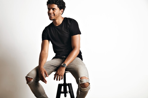 Free photo playful smiling black model in grey jeans and plain cotton black t-shirt on stool isolated on white