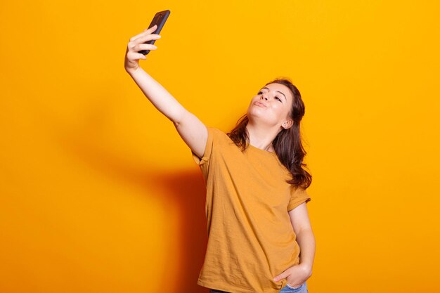 Playful woman holding mobile phone to take selfies in studio. Caucasian person using smartphone with technology, taking pictures over orange background. Happy adult with modern device