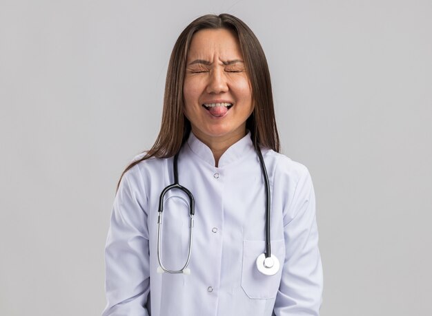 Playful young asian female doctor wearing medical robe and stethoscope showing tongue with closed eyes isolated on white wall