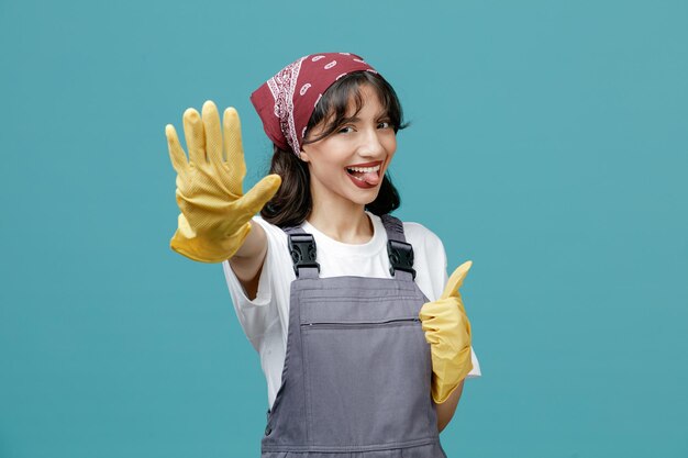 Playful young female cleaner wearing uniform bandana and rubber gloves looking at camera showing tongue and thumb up and making stop gesture isolated on blue background