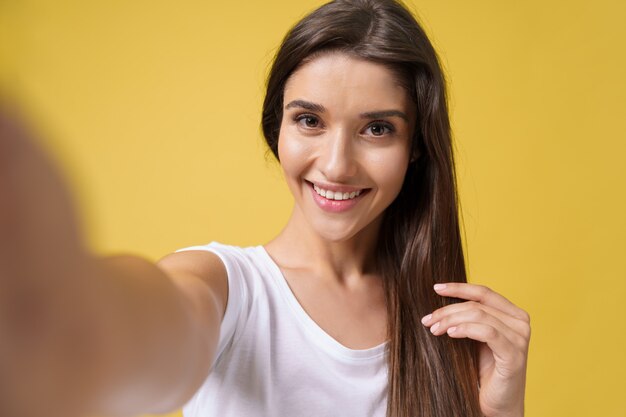 Pleasant attractive girl making selfie in studio and laughing. Good-looking young woman with brown hair taking picture of herself on bright yellow background.