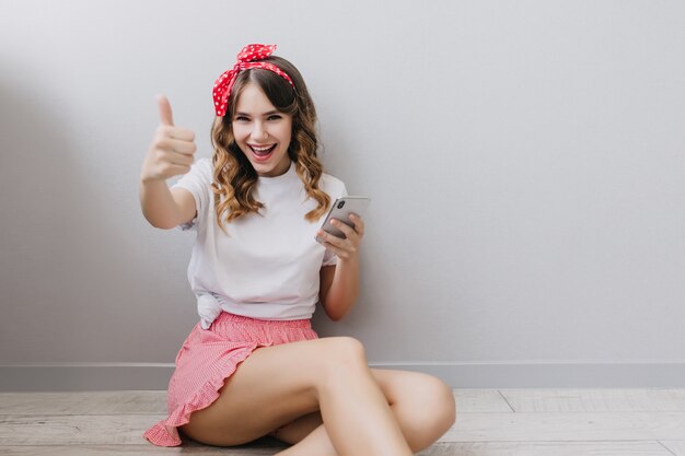 Pleasant caucasian girl with red ribbon in dark hair sitting on the floor at home. Indoor shot of smiling female model in pink shorts.