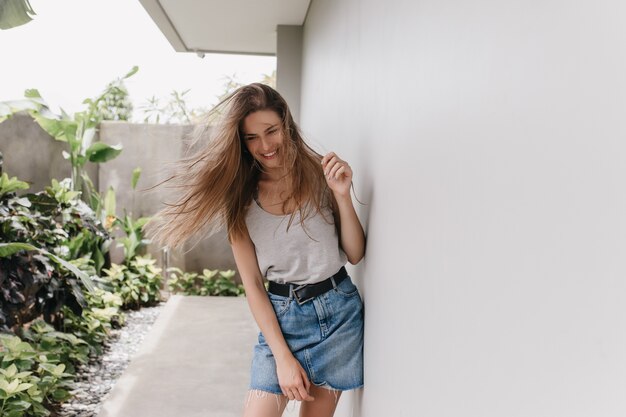 Pleasant girl posing with long hair waving and laughing beside white wall. Cheerful female model in denim skirt spending time outdoor in warm day.