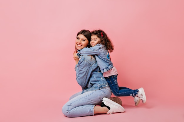 Free photo pleasant kid embracing mother on pink background. studio shot of blissful mom and little daughter in jeans.