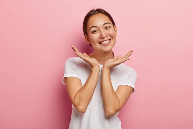 Pleasant looking Asian female model smiles gladfully, spreads palms near face, expresses positive emotions, wears white t shirt, has appealing appearance, healthy skin, isolated on pink  wall