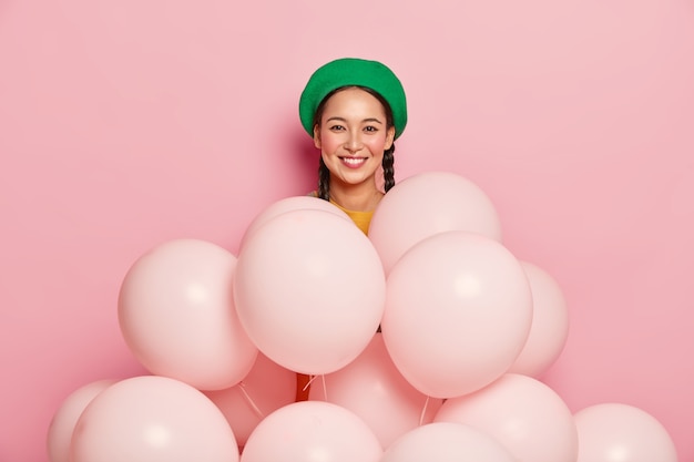 Pleasant looking Asian female model wears green beret, stands near many balloons, poses over pink background, celebrates birthday