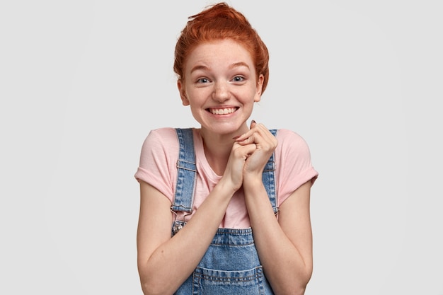 Free photo pleasant looking joyful red head young lady with glad facial expression, keeps hands together, has blue eyes full of happiness, stands against white wall, recieves nice gift