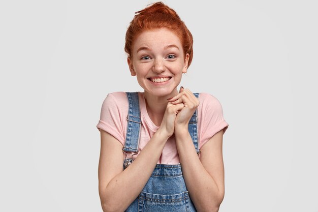 pleasant looking joyful red head young lady with glad facial expression, keeps hands together, has blue eyes full of happiness, stands against white wall, recieves nice gift