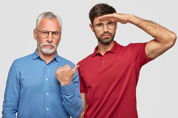 Free photo pleasant looking successful bearded old businessman in elegant-shirt indicates with thumb at his son who keeps hand near forehead and looks attentively into distance, explains about family business