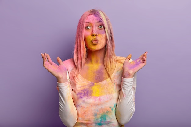 Pleasant looking woman has folded lips, spreads hands, wears casual clothes, has glad expression, dirty with colorful powder, isolated over lilac wall. Colored dust explode