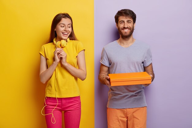 Pleasant looking woman keeps hands in anticipation, looks curiously at cardboard box, being interesting what is inside. Happy young man holds little contrainer or package, gives present to girlfriend