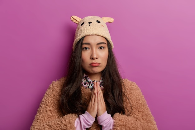 Please, forgive me. Sad mixed race woman begs for help, keeps palms in praying gesture, wears funny teenage hat, winter coat