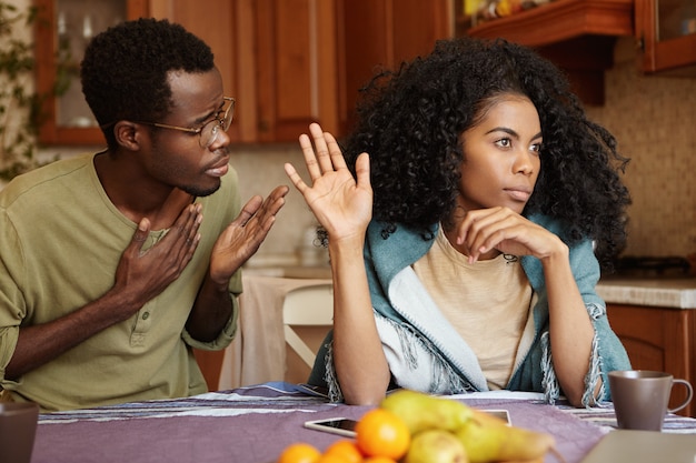 Please forgive me. Unhappy African American male cheater holding hand on his chest apologizing to beautiful indifferent woman who ignoring and refusing all his excuses, telling him to get lost