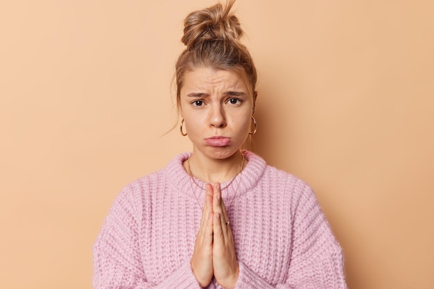 Free photo please help me. upset young woman with combed hair purses lis looks sadly at camera keeps palms pressed together begs you asks for forgiveness wears casual knitted sweater isolated on beige background