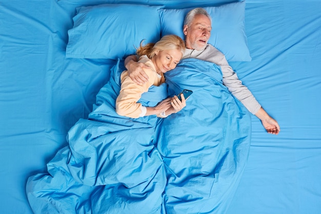 Pleased addicted to modern technologies woman uses mobile phone in bed receives hug from sleeping husband scrolls internet before sleep. Middled aged family couple have rest at cozy bedroom.