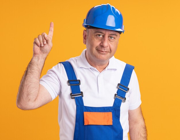 Pleased adult builder man in uniform points up isolated on orange wall