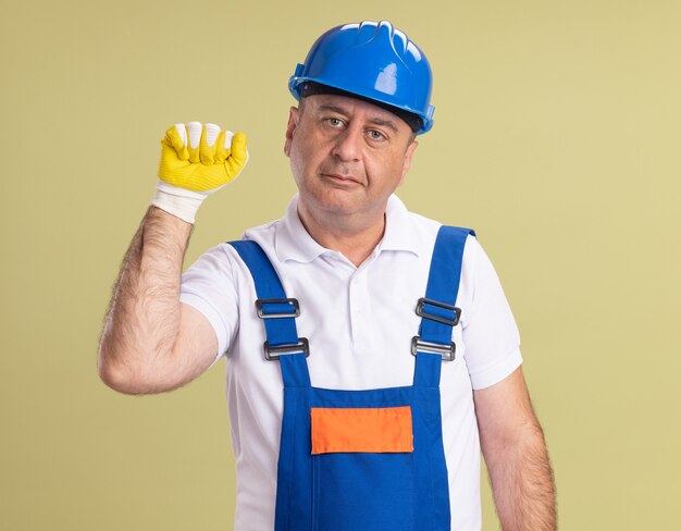 Pleased adult builder man in uniform wearing protective gloves keeps fist up isolated on olive green wall