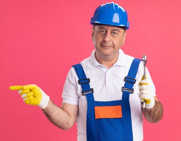 Pleased adult caucasian builder man in uniform wearing protective gloves holds wrench and points at side on pink