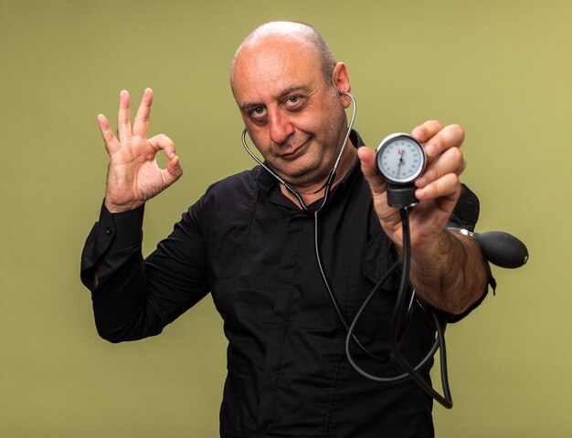 pleased adult ill caucasian man holding sphygmomanometer and gesturing ok sign isolated on olive green wall with copy space