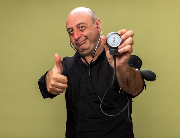 pleased adult ill caucasian man holding sphygmomanometer and thumbing up isolated on olive green wall with copy space