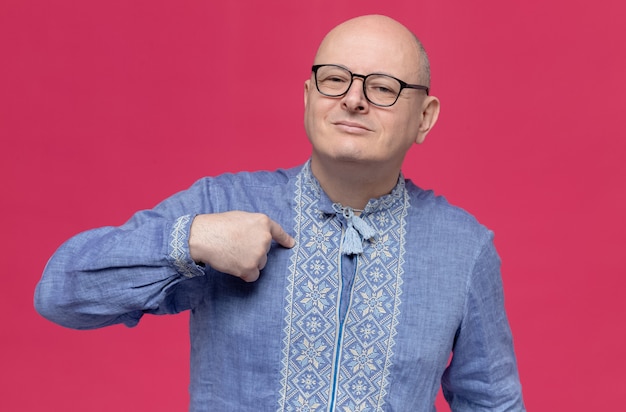 Pleased adult man in blue shirt wearing glasses pointing at himself 