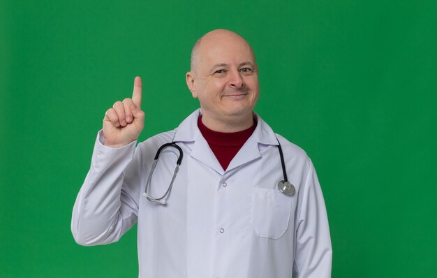 Pleased adult slavic man in doctor uniform with stethoscope pointing up