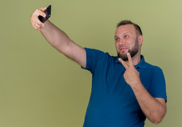 Pleased adult slavic man doing peace sign and taking selfie 