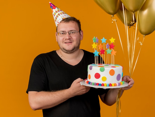 Pleased adult slavic man in optical glasses wearing birthday cap holds helium balloons and birthday cake looking at camera 