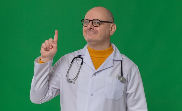 Pleased adult slavic man with optical glasses in doctor uniform with stethoscope pointing up