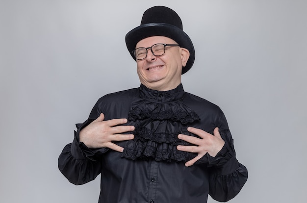 Free photo pleased adult slavic man with top hat and optical glasses in black gothic shirt putting hands on his chest