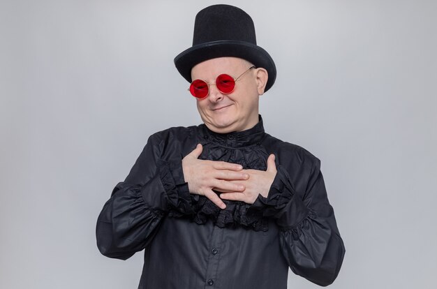 Pleased adult slavic man with top hat and with sunglasses in black gothic shirt putting hands on his chest 