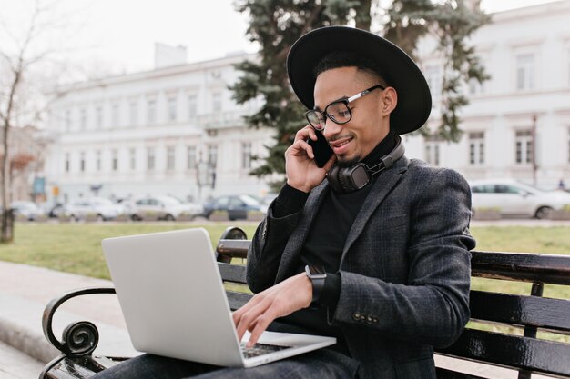 Pleased african freelancer talking on phone and typing on keyboard. Outdoor photo of international student in black outfit using laptop on nature.