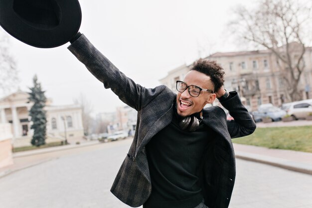 Pleased african man enjoying life in park. Outdoor portrait of laughing black guy waving hat on blur town.